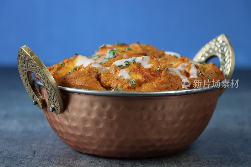 卡扎伊(kadhai)烹饪锅/上菜碗的特写，里面装满了自制的沙希奶酪(咖喱奶酪)，浓汁的奶油，西红柿，印度香料，灰色的石板表面，蓝色的背景，聚焦于前景